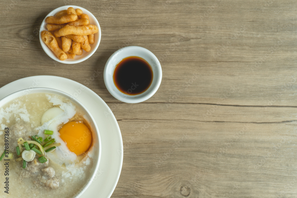 Close up view of rice porridge or congee with minced pork, egg, slice ginger and slice scallion topping near by deep-fried dough stick and soy sauce in a white bowl on wooden desk. Breakfast concept. Close up view of rice porridge or congee with minced pork, egg, slice ginger and slice scallion topping near by deep-fried dough stick and soy sauce in a white bowl on wooden desk. Breakfast concept.