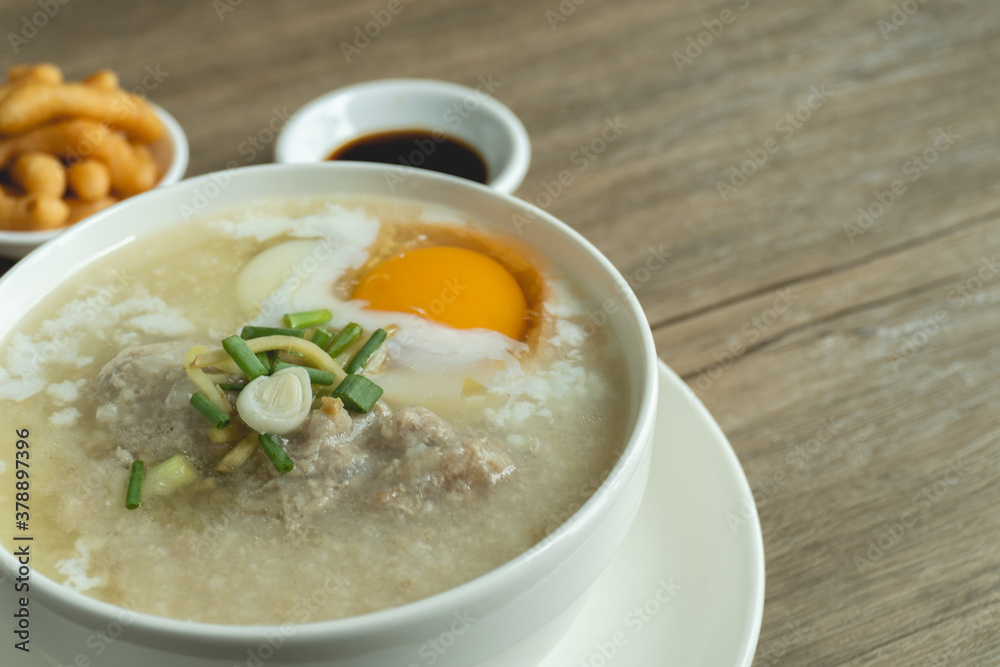 Close up view of rice porridge or congee with minced pork, egg, slice ginger and slice scallion topping near by deep-fried dough stick and soy sauce in a white bowl on wooden desk. Breakfast concept. Close up view of rice porridge or congee with minced pork, egg, slice ginger and slice scallion topping near by deep-fried dough stick and soy sauce in a white bowl on wooden desk. Breakfast concept.