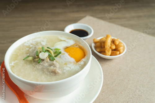 Close up view of rice porridge or congee with minced pork, egg, slice ginger and slice scallion topping near by deep-fried dough stick and soy sauce in a white bowl on wooden desk. Breakfast concept.