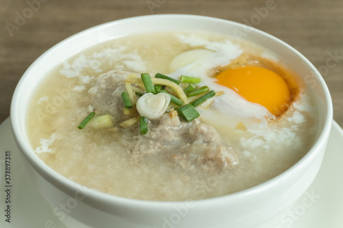 Close up view of rice porridge or congee with minced pork, egg, slice ginger and slice scallion topping in a white bowl on wooden desk. Breakfast concept.
