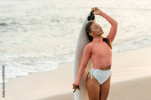 Young attractive surfer woman with white board at sunset on the ocean. Bali Indonesia. Summer time, sports, travel content.