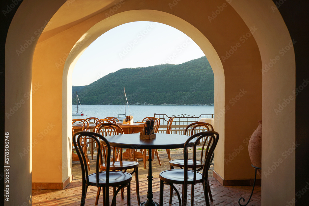 view from the street with tables and chairs to a mountain lake, sailboats and mountains at sunset
