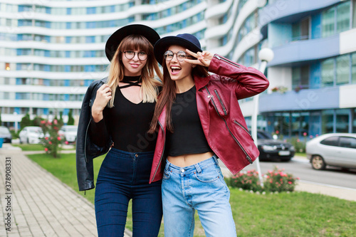 Two carefree smiling women posing  over modern city background. Wearing wool hat , leather jacket and jeans. Friends hugging. Happy emotions . © Svetlana Sokolova