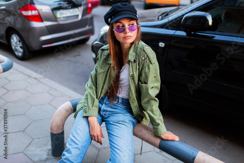 Fashionable woman with long brunet hairs posing outdoors in big city near road . Wearing stylish green jacket , black cap and purple sunglasses. © Svetlana Sokolova