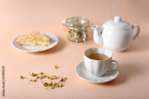 White cup with brewed chamomile, teapot, pie and jar of tea on a pink background.