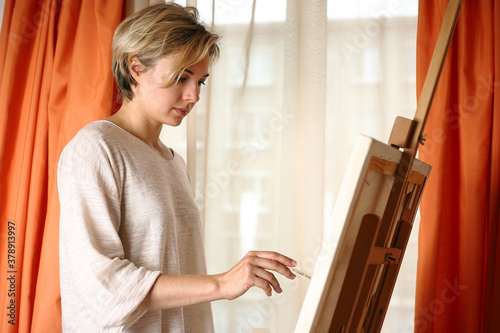 Young blonde caucasian woman with easel in a white sweater draws on the background of the window