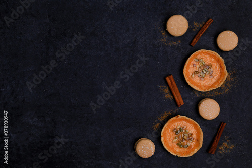 autumn orange creamy pumpkin pies for halloween with seeds, cinnamon sticks, several macaroon cakes on a black background, top view, place for text, flat lay