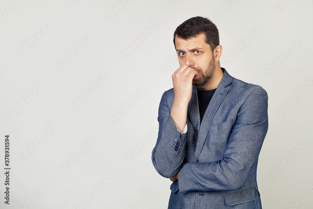 Young businessman with a smile, a man with a beard in a jacket, looks tense and nervous with hands on his lips, biting his nails. Anxiety problem. Portrait of a man on a gray background Young businessman with a smile, a man with a beard in a jacket, looks tense and nervous with hands on his lips, biting his nails. Anxiety problem. Portrait of a man on a gray background