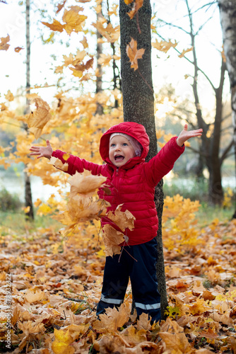 A girl in a red jacket and dark pants throws bright foliage up. The foliage falls and the child rejoices and laughs. Around  autumn colorful foliage covers the ground. Around the trees. a park
