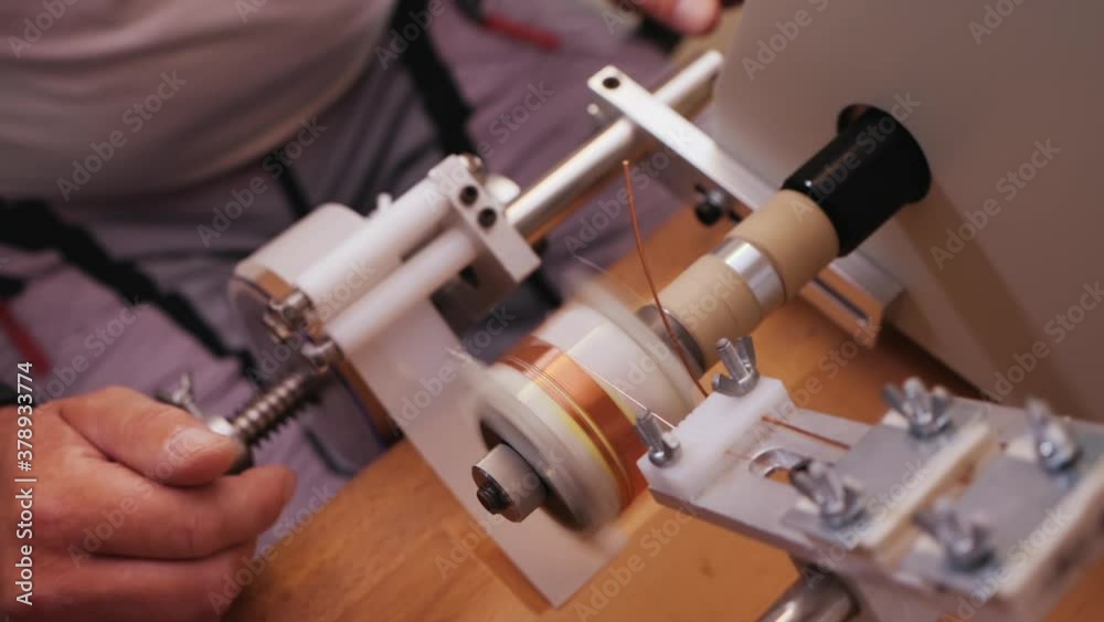 Hands operate coil winding machine, making a transformer component - reeling conducting wire on plastic support, close up, slow motion, top view