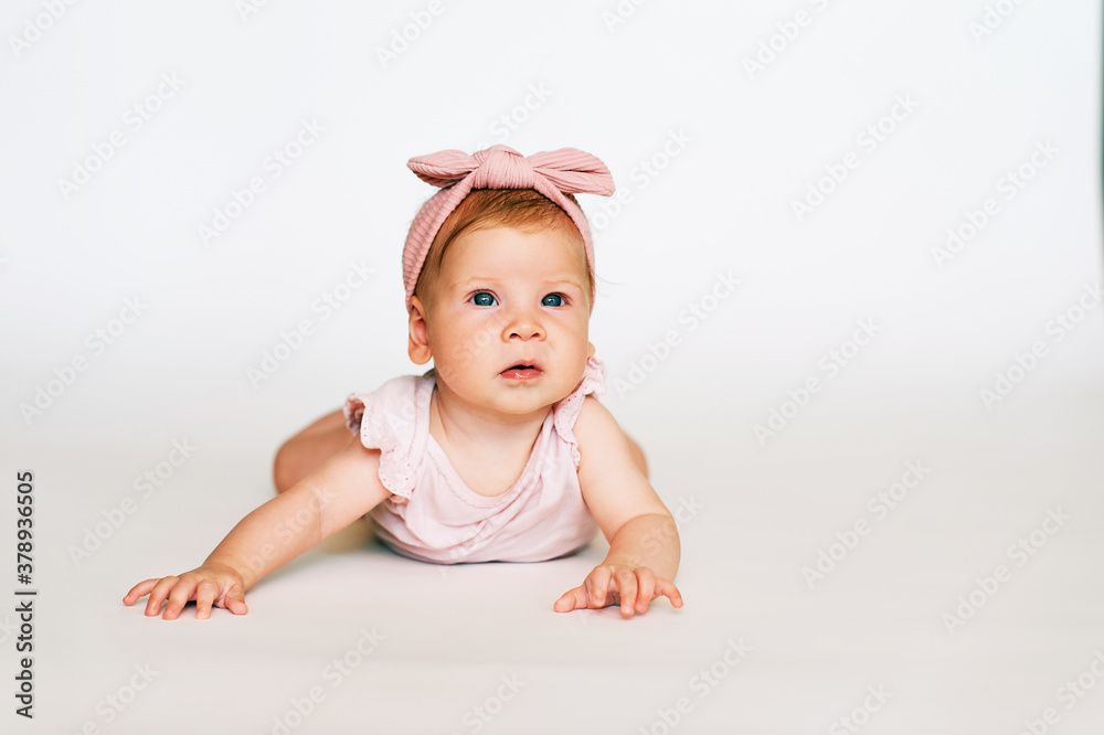 Portrait of adorable red-haired baby lying on belly, white background, wearing pink body and headband, looking up Portrait of adorable red-haired baby lying on belly, white background, wearing pink body and headband, looking up