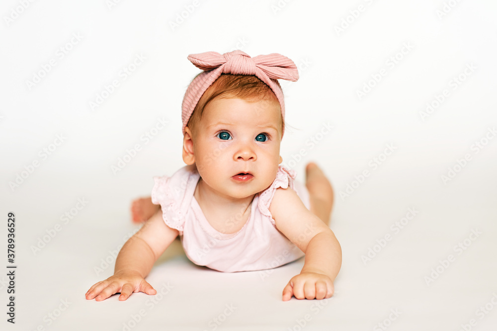 Portrait of adorable red-haired baby lying on belly, white background, wearing pink body and headband Portrait of adorable red-haired baby lying on belly, white background, wearing pink body and headband