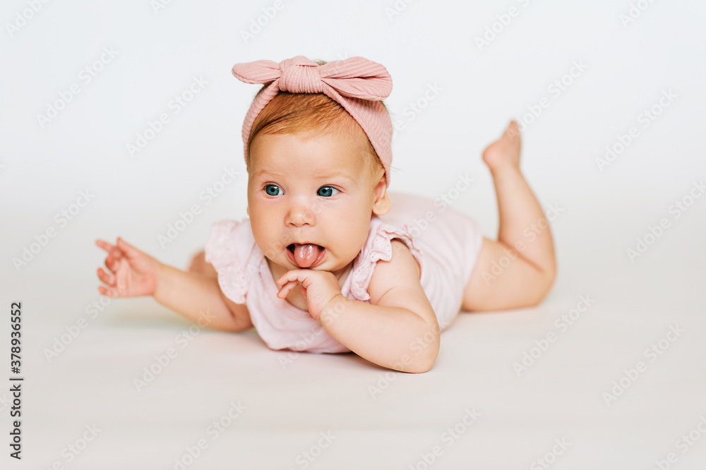 Portrait of adorable red-haired baby lying on belly, white background, wearing pink body and headband, licking hand Portrait of adorable red-haired baby lying on belly, white background, wearing pink body and headband, licking hand