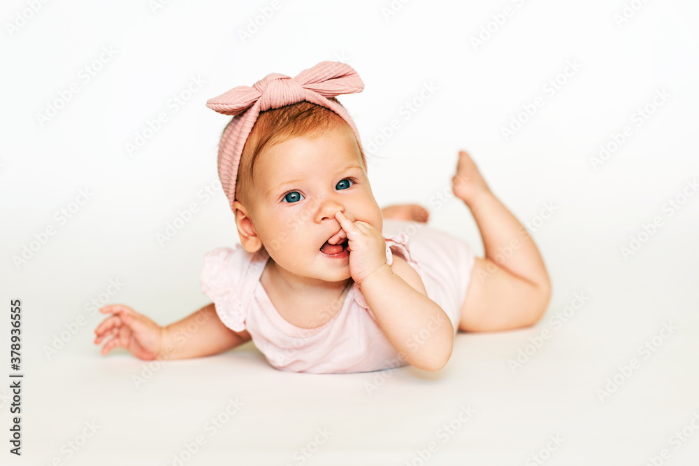 Portrait of adorable red-haired baby lying on belly, white background, wearing pink body and headband Portrait of adorable red-haired baby lying on belly, white background, wearing pink body and headband