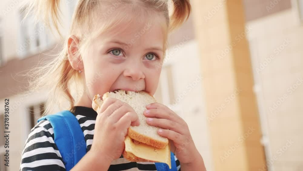 schoolgirl eating a sandwich during recess in school. kids education concept. lifestyle little girl kid schoolgirl with a backpack eating a sandwich. kid child is having lunch at school schoolgirl eating a sandwich during recess in school. kids education concept. lifestyle little girl kid schoolgirl with a backpack eating a sandwich. kid child is having lunch at school