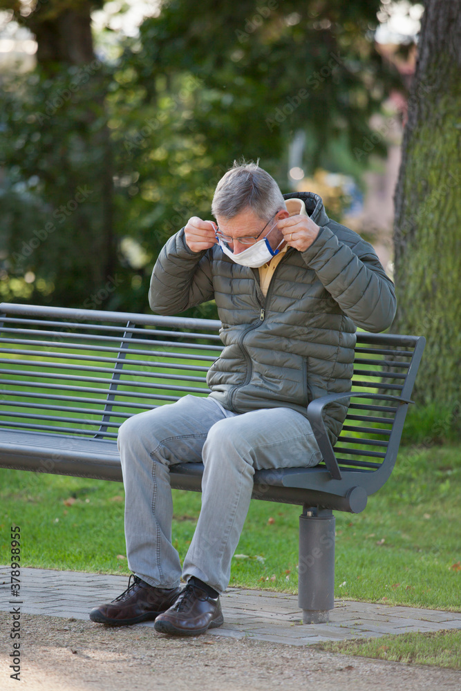 Mature man puts a face mask on or off while sitting on a bech in a park Mature man puts a face mask on or off while sitting on a bech in a park