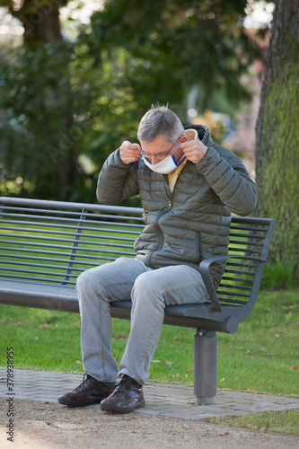 Mature man puts a face mask on or off while sitting on a bech in a park