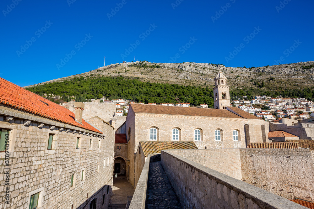 Colorful fortress street walk scene, clear sky sunny day. Church front and building roofs. Winter view of Mediterranean old city of Dubrovnik, famous European travel and historic destination, Croatia