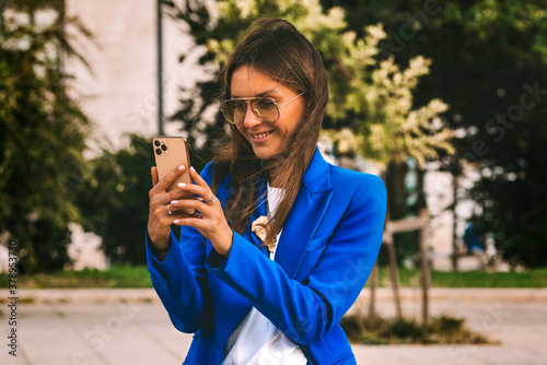 Happy smiling businesswoman in blue blazer using smartphone. Video call. Talking with friends on cell phone. Typing message or shop online or checking mobile application or social media
