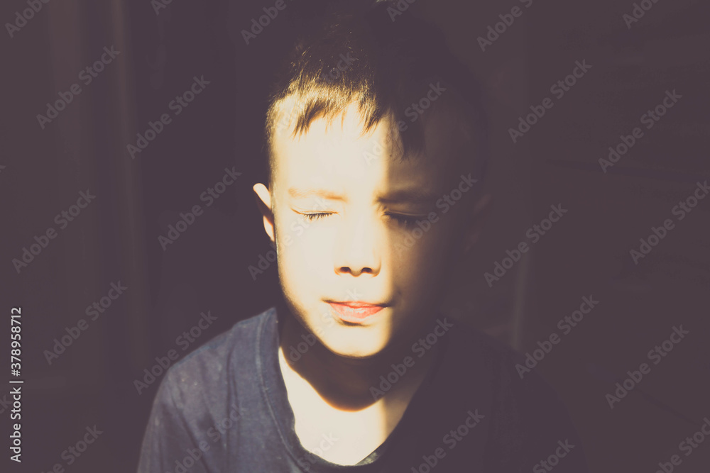 A young European boy squints in the bright sunlight from the window A young European boy squints in the bright sunlight from the window