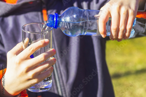 Pour clear cold water into a glass glass on the street against the background of green trees and grass.