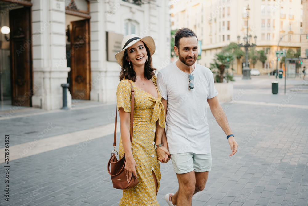 A happy girl in a hat and a yellow dress with a plunging neckline and her boyfriend with a beard and sunglasses are walking holding each other's hand in the old town. A couple of tourists in Valencia.