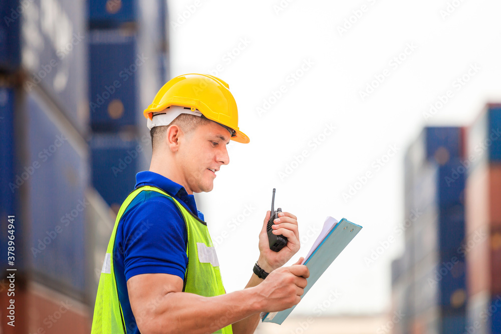 Foreman worker in hard hat and safety vest holding clipboard checklist and talks on two-way radio at containers cargo Foreman worker in hard hat and safety vest holding clipboard checklist and talks on two-way radio at containers cargo