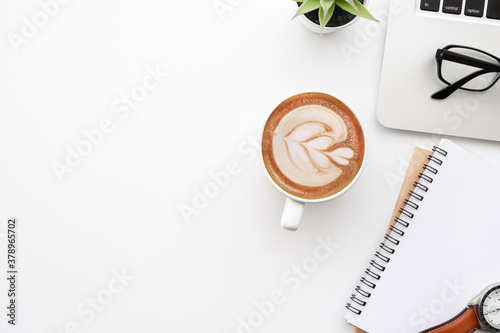 White office desk table with blank notebook, laptop computer, cup of coffee and supplies. Top view with copy space, flat lay.