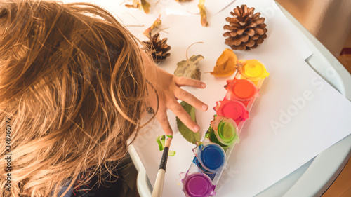 Toddler boy is home schooled paiting yellow leaves and pine cones in fall with eco friendly paint. Sensorial activity at home.