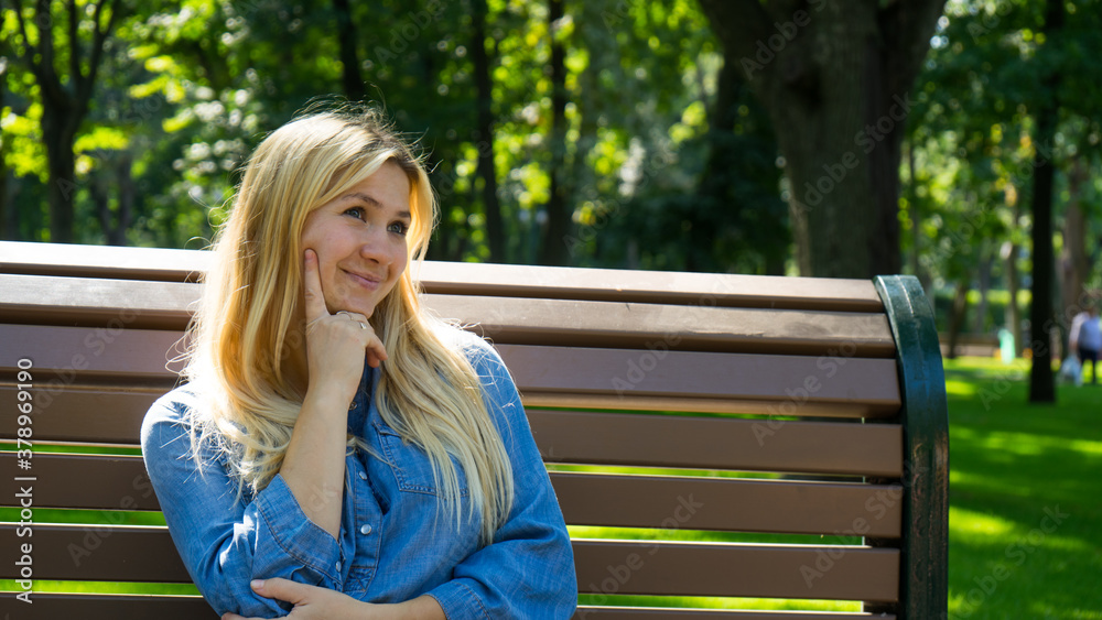 Portrait of attractive casual blond girl thoughtfully looking away and thinking on something sitting in the sunny city park. Pretty young thoughtful woman confused about something sits on bench Portrait of attractive casual blond girl thoughtfully looking away and thinking on something sitting in the sunny city park. Pretty young thoughtful woman confused about something sits on bench