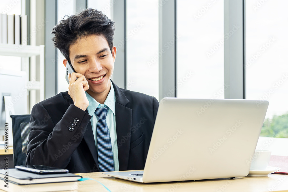 Smiling young asian businessman sitting at desk talking on mobile phone and using laptop in workplace Smiling young asian businessman sitting at desk talking on mobile phone and using laptop in workplace