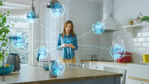 Internet of Things Concept: Young Woman Using Smartphone in Kitchen. She controls her Kitchen Appliances with IOT. Graphics Showing Digitalization Visualization of Connected Home Electronics Devices photo