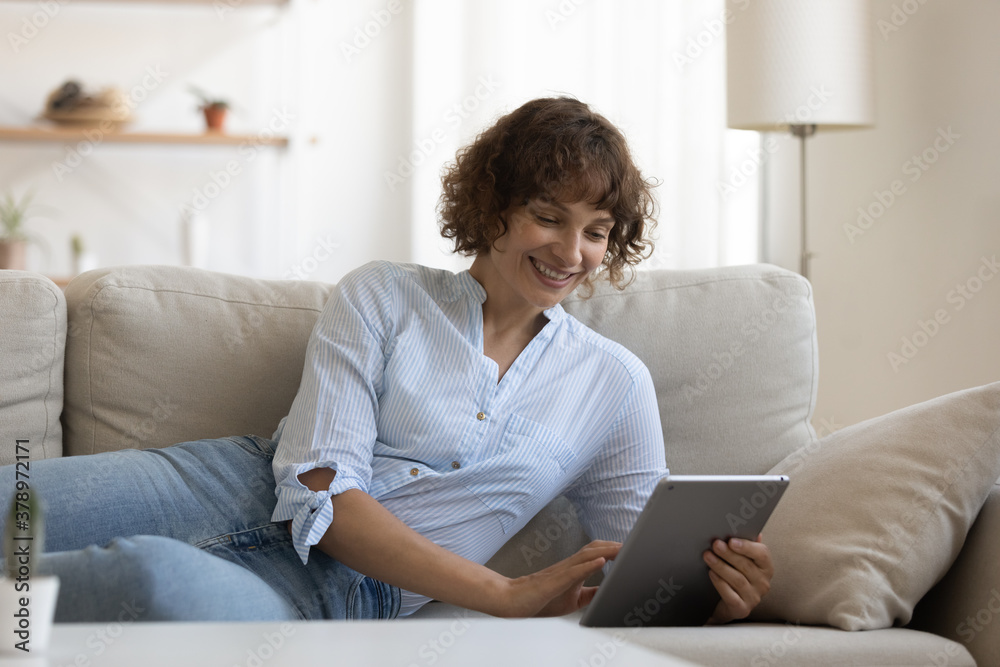 Smiling young Caucasian woman sit rest on couch in living room talk on video call on tablet gadget. Happy female relax on sofa at home browse use internet, have webcam virtual conversation. Smiling young Caucasian woman sit rest on couch in living room talk on video call on tablet gadget. Happy female relax on sofa at home browse use internet, have webcam virtual conversation.