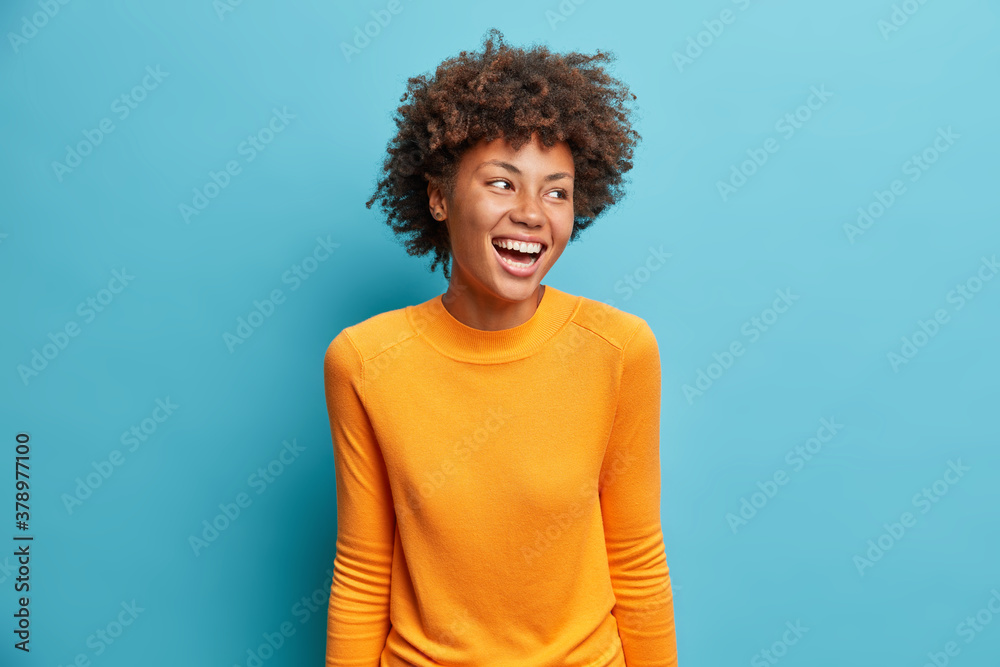 Positive cheerful young African American woman laughs positively and looks aside concentrated happily dressed in casual orange jumper isolated on blue studio background. People emotions concept Positive cheerful young African American woman laughs positively and looks aside concentrated happily dressed in casual orange jumper isolated on blue studio background. People emotions concept