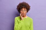 Emotional curly haired lady cannot believe in shocking news stares through round glasses wears knitted green jumper isolated on purple background. Surprised Afro American woman expresses amazement