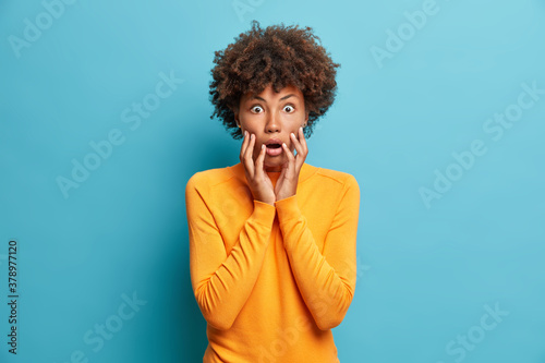 Scared dark skinned young woman stares bugged eyes and holds face keeps mouth opened wears casual jumper poses indoor. Frightened Afro American girl gazes scaredly at something embarrassing.