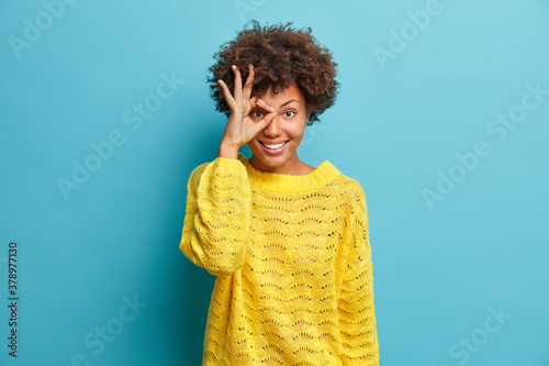 Positive dark skinned woman makes okay gesture over eye and smiles broadly wears knitted yellow sweater. Happy attractive female with Afro hair feels glad likes your idea isolated on blue wall