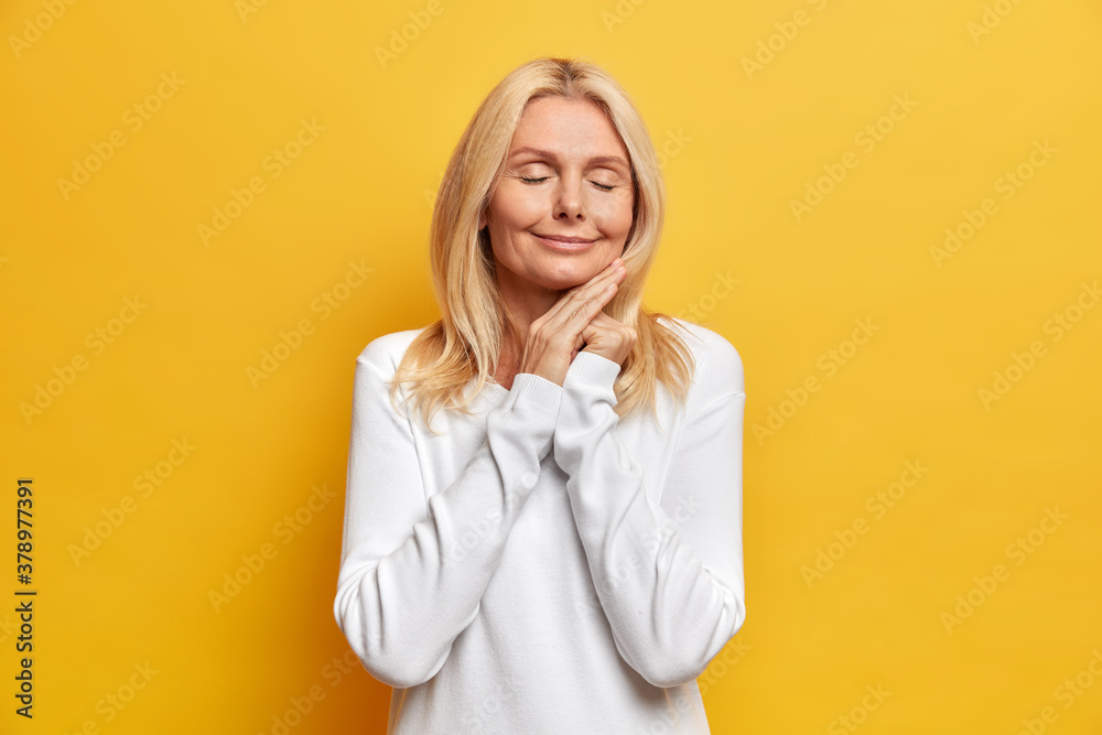 Aging and beauty concept. Charming senior wrinkled Caucasian woman touches face gently stands with closed eyes smiles tenderly enjoys pleasant moment in life isolated on yellow studio background. Aging and beauty concept. Charming senior wrinkled Caucasian woman touches face gently stands with closed eyes smiles tenderly enjoys pleasant moment in life isolated on yellow studio background.