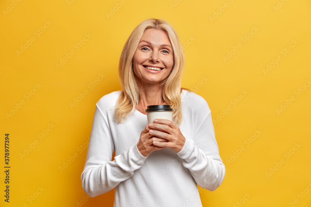Isolated shot of cheerful adorable woman of middle age woman has lively happy talk while drinks coffee expresses positive emotions takes break dressed in white sweater poses against yellow background Isolated shot of cheerful adorable woman of middle age woman has lively happy talk while drinks coffee expresses positive emotions takes break dressed in white sweater poses against yellow background