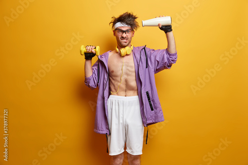 Photo of displeased sporty man raises heavy dumbbell and bottle with strength power, builds body in fitness gym and wears sport clothes. Determined guy has weight training, listens music in headphones