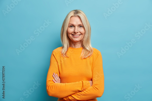 Portrait of good looking blonde woman stands self assured against blue studio background smiles gently and keeps arms folded glad to meet old friend. People age beauty sincere emotions concept