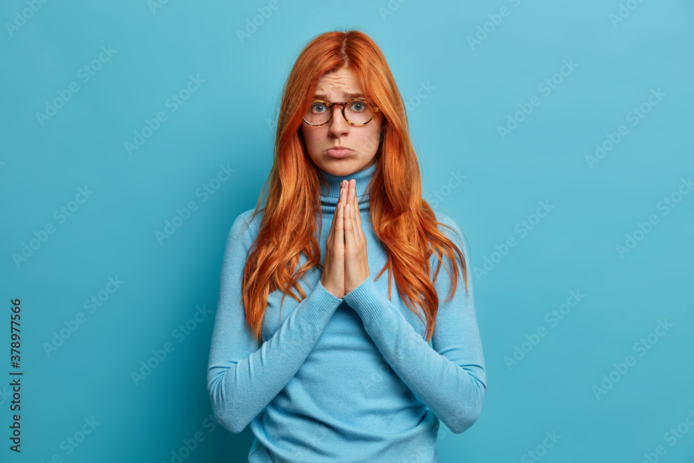 Sad pitty woman looks with imploring expression at camera presses palms together asks for favor and needs help poses against blue background. Redhead girl has big hope to get something desirable