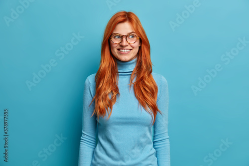 Portrait of cheerful ginger millennial girl wears casual turtleneck round spectacles, feels joyful as looks aside and poses against blue studio background. Positive emotions face expressions concept