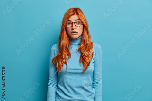 Portrait of amazed redhead woman feels very shocked poses with opened mouth and holds breath from amazement. Beautiful speechless lady in eyeglasses looks in disbelief poses over blue studio wall