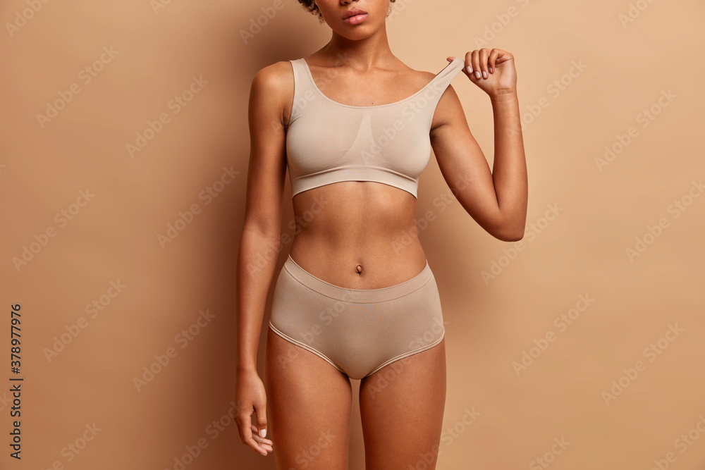 Fototapeta premium Femine beauty lines concept. Slim woman with dark skin flat stomach wears high waist panties and bra. Female model poses in underwear against brown background