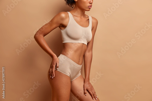 Body care and beauty concept. Unrecognizable dark skinned woman in underwear stands against brown studio background. Healthy breast
