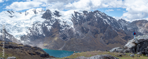 Panorama landscape of man standing in a rock during hike/trail with huge mountains with snow and a lake with blue sky and clouds, in cordilera Huaytapallana, Huancayo, peru