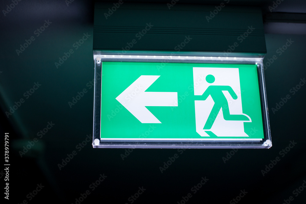 Green and white exit sign hangs on the ceiling of the corridor, lighted in the darkness. Arrow points to the left. Green and white exit sign hangs on the ceiling of the corridor, lighted in the darkness. Arrow points to the left.