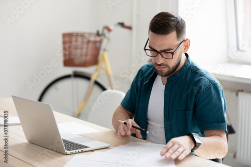 Serious young businessman wearing glasses working with documents, sitting at work desk with laptop in modern office, busy focused worker entrepreneur making notes, online research, financial report