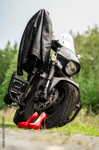 Against the background of a green forest, there is a black motorcycle, a leather jacket hangs on it, and red shoes are pulled off next to it